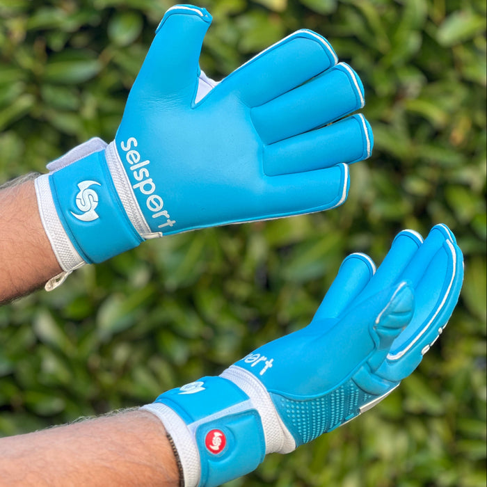 Blue Selsport goalkeeper gloves with white accents on a person's hands against a green leafy background
