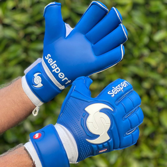 Blue Selsport goalkeeper gloves with white logos held by a person against a green hedge background
