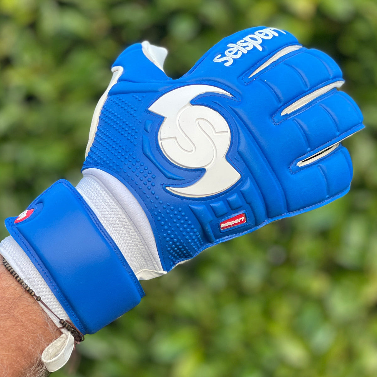 Blue Selsport goalkeeper glove with a white logo on a blurred green background