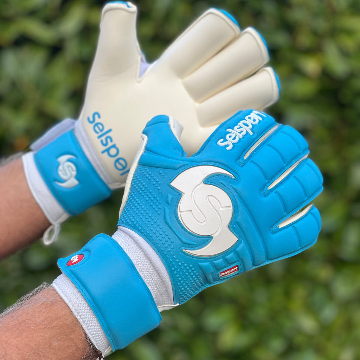 Cyan Selsport goalkeeper gloves with a visible brand logo on a blurred green background