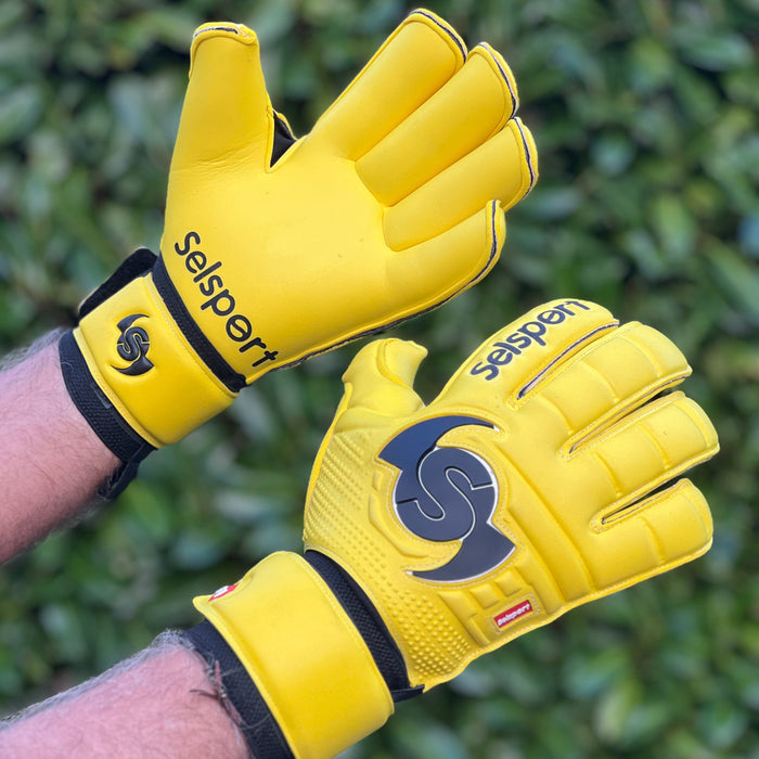 Yellow Selsport goalkeeper gloves with a logo held against a green leafy background