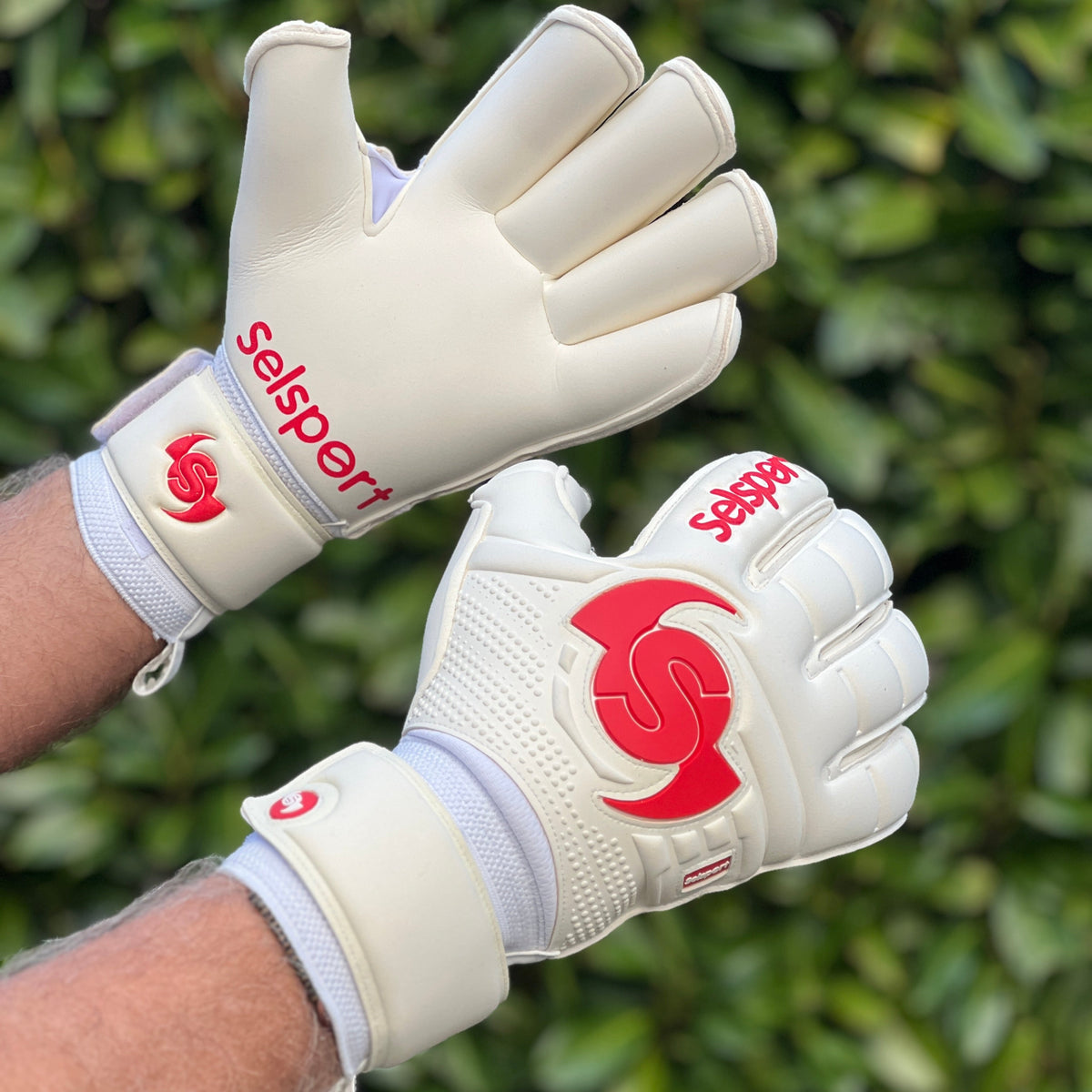 White Selsport goalkeeper gloves with red logo worn by a person against a green leafy background
