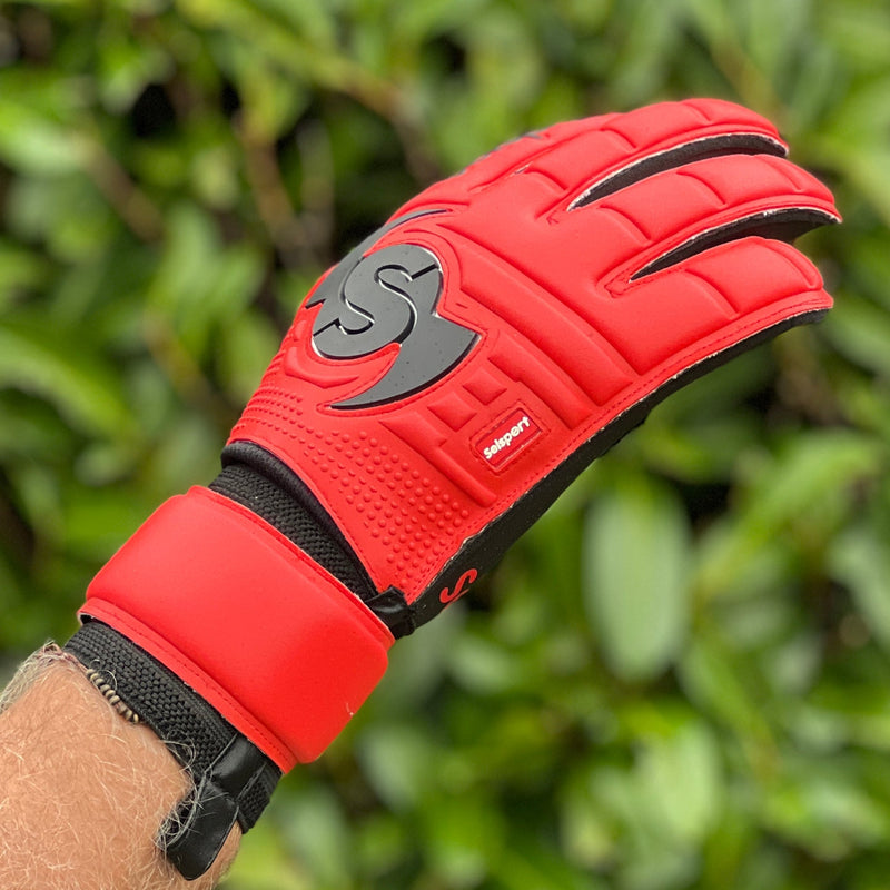 Red Selsport goalkeeper glove with black logo on a blurred green background