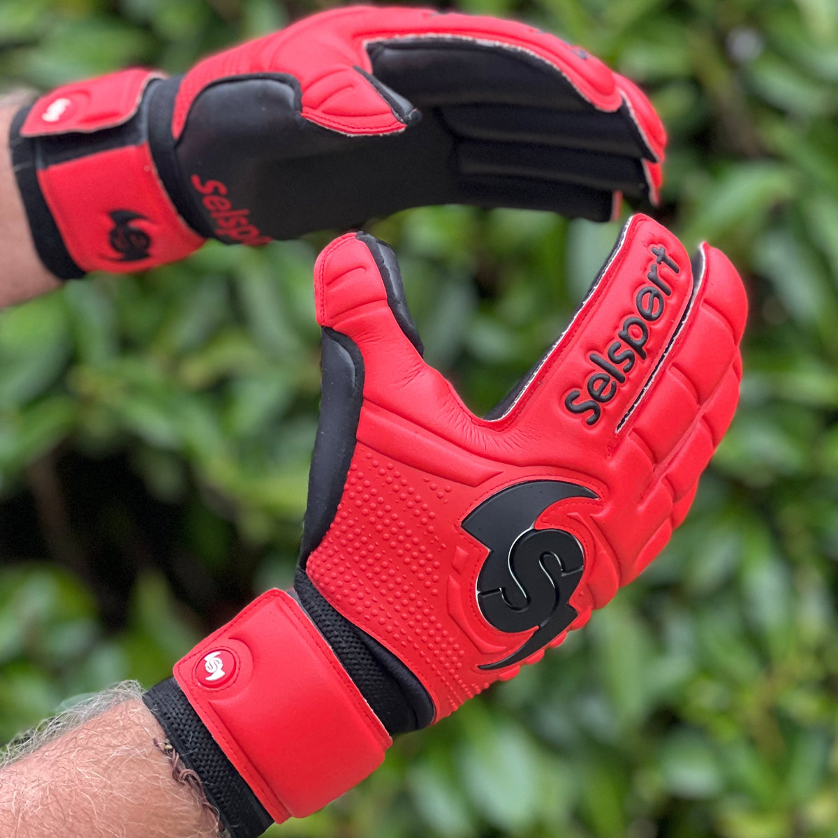 Red Selsport  goalkeeper gloves with black accents on a blurred green background