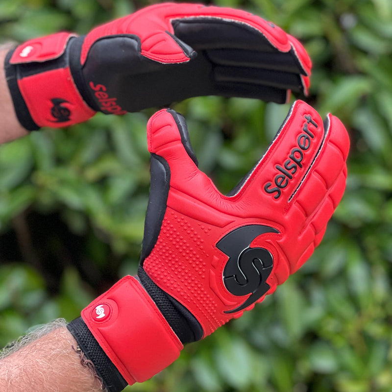 Red Selsport  goalkeeper gloves with black accents on a blurred green background
