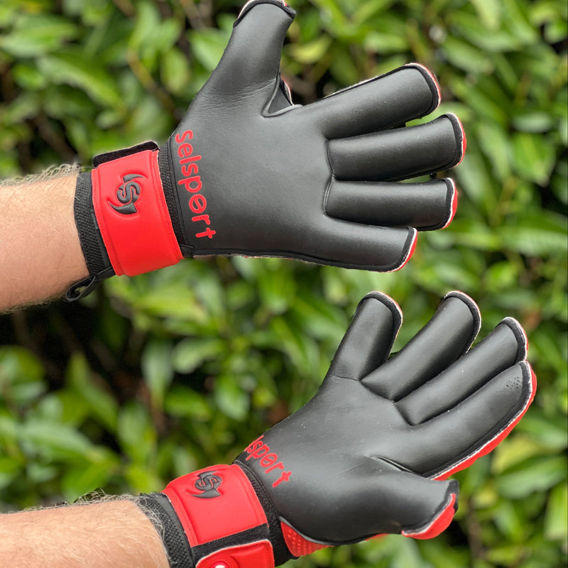Black and red Selsport goalkeeper gloves with visible branding against a green leafy background