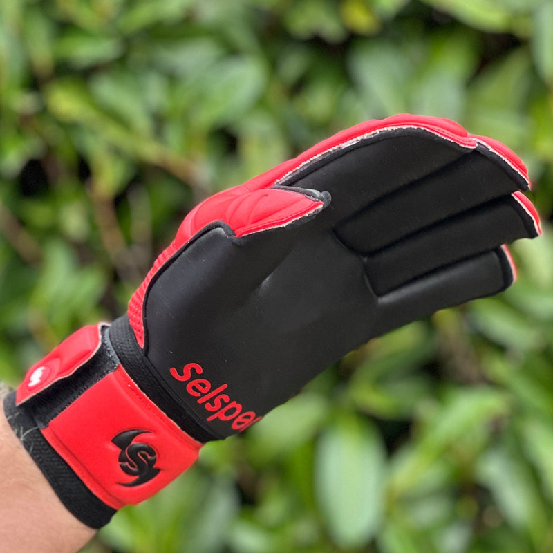 Red and black goalkeeping glove with 'Selsport' branding against a green leafy background